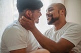 Two men wearing white tee-shirts embrace and lean in for a kiss.