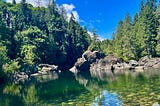 A rocky and treed area surrounds a flowing river. It is shallow with rocks on the bottom. A blue sky is overhead