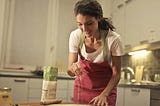 woman cooks and adds spice in kitchen. She is smiling, and wearing a red apron