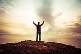 This is a man who is standing on a rock. The colour of the rock is dark brown. The man is standing in front of the sea. We can see clouds in the background. The man is raising his hands in front of the sky. It seems he is admiring the beauty of nature.