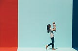 A woman with long brown hair holds up a baby in front of a colourful red and blue street