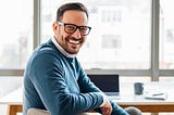 A writer smiling while sitting at a wooden desk. In the background, a laptop, coffee mug, and notebook are visible.