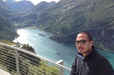 A picture of the author with a view of the blue water of the fjord and green mountains in the background