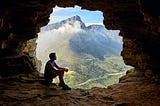 This is a picture of a man sitting alone in a cave. He is looking outside the cave towards the sky. The colour of the cave is dark brown. We can also see hills in the background partially covered with clouds. The cave is situated at a considerable height.