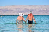 Older man and woman holding hands and walking into the sea.