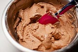 A red spatula is resting in the chocolate batter in a metal baking bowl