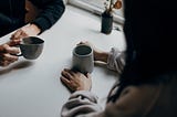 Two people in conversation, sitting across the table and holding ceramic coffee mugs