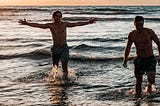 Two men in swim trunks standing in the surf.