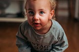 This is a baby child. He is wearing a grey T-shirt. His eyes are light blue. His complexion is white. His hair is golden brown. This child appears to be highly sensitive.