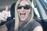 Photo of Author giving the thumbs up from a selfie taken inside the car, wearing sunglasses and a grey t-shirt and with blonde hair just past her shoulders.