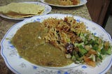 White plate in foreground shows a brown curry with chopped cucumber, onion and tomato salad and brown rice on either side. Another plate has a couple of roasted papads (lentil crisps). A third plate in the background has the same dishes as the first one.