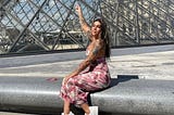 Woman in a pink flowery dress sits on a polished concrete ledge pointing up to the Louvre pyramid.