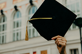 A hand holds a graduation cap in the air, in front of some lovely, old looking building.