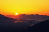 The sun is about to set behind a mountain chain, colouring the sky golden and the mountain ridges red. There is flat land with several lakes coloured gold and red in the mid-section of the photo. Fog coloured slightly blue whiffs over the earth.