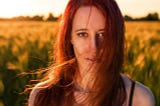 A woman with long, red hair and a sad expression standing in front of a wheat field.