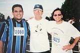 Three men standing side by side at an outdoor event, smiling at the camera. The man on the left wears a blue and black striped sports jersey with a race number ‘1060,’ and the man on the right wears sunglasses and a white shirt with a race number ‘1025.’ The man in the center wears a white T-shirt with a hiking graphic and a ‘Grand Canyon’ hat.