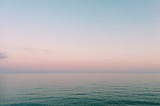 A beach at sunrise, with pastel hues in the sky and on the water blurring the transition between them.