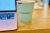 “Tiffany & Company Coffee Cup in Bone China, Everyday Objects” stained on the rim by red lipstick, next to an open laptop, on top of a wooden desk