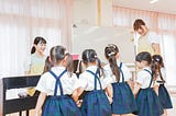 Young children in school uniforms standing in front of a piano, receiving music instruction from a teacher while another teacher smiles nearby. The children have their backs to the camera, and the classroom has light-colored curtains and a whiteboard