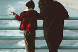 young boy standing next to his mother, overlooking the sea from a railing, viewed from the back.