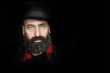 Sophisticated, rather vain-looking bearded man staring confidently, wearing hat and red scarf, background black