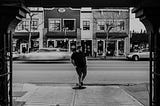 A black and white photograph of a man standing on the sidewalk, waiting to cross a street. The background features a row of small shops and buildings. A car is seen driving by in a blur, capturing motion, while the man remains still. The scene appears to be from a small town or suburban area.