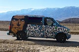 Painted van stopped on the side of a road. there is a man’s face painted near the back of the van with the words “Rock and Roll can Never Die” on the side. Mountains are visible in the distance.