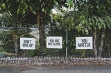 A chain-link fence with three signs that read ‘DON’T GIVE UP,’ ‘YOU ARE NOT ALONE,’ and ‘YOU MATTER,’ surrounded by greenery and trees in a peaceful outdoor setting.