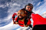 Two skydivers in tandem, the foreground skydiver grinning widely, as they freefall against a backdrop of bright blue sky and soft white clouds, embodying the thrill of skydiving.