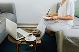 woman sits on a couch writing something in a notebook with a laptop and coffee on a small table in front of her.