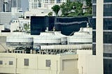 View of rooftop cooling towers and HVAC systems on top of a building in a cityscape, with high-rise buildings and trees visible in the background.