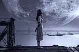 Woman standing by a shore in the dark, looking at the moon and clouds.