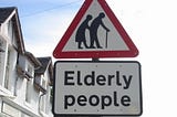 A British road sign, a red triangle with two people hunched over using walking sticks, with a rectangular sign below with the words, “Elderly people”.