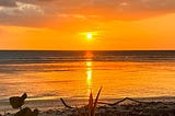 Tobago beach sunset with the sun shimmering over the sea as the central focus of the photo. Lots of oranges and greys.