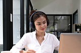 A woman is seated at a desk, wearing headphones and working on a laptop. She holds a pencil and writes in a notebook, with eyeglasses placed on the desk beside a coffee cup. The setting is an office or home workspace with natural light coming in from the background.