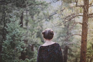 A young woman stands in the forest. We see her back as she looks forward.