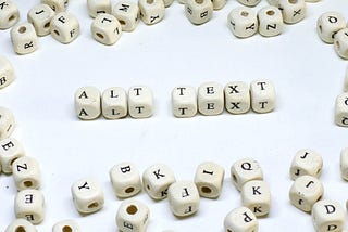 A group of alphabetic dice spelling out the words “ALT TEXT”. They are on a white surface and surrounded on all sides by perhaps two dozen similar dice that have random letters that spell nothing. Each die has a small hole going through the center; they’re the type of dice you see used to make bracelets that spell out messages