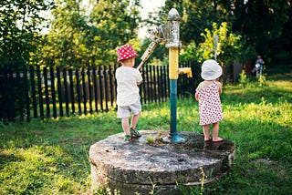 Two young children, wearing sun hats, stand by an outdoor water pump in a grassy area on a sunny day. One child is operating the pump handle while the other watches. The scene is peaceful, surrounded by greenery and a wooden fence in the background.