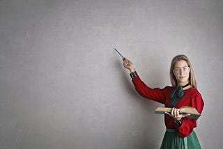 photo entitled “Woman in Discussing A Lesson Plan.” she is dressed conservatively, holding a large open book in her left hand. she is holding a pointer in her right hand, which gestures at what appears to be a large blackboard where all the previous writings/markings have been erased.