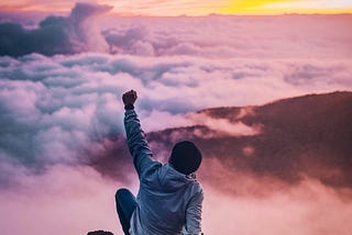 male sitting on mountaintop above the clouds, with fist raised triumphantly, gazing into sunrise