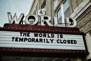 Marquee of an old theater with the word ‘WORLD’ in large letters on top. The sign below reads ‘The world is temporarily closed,’ referencing the global impact of the COVID-19 pandemic. The building has a classic, vintage aesthetic.