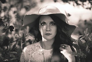 Head shot of a dark-haired woman among trees in a floppy hat with wide-eyed, eerily angry expression.