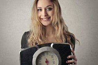 A smiling young woman holds up an old-style analog scale.