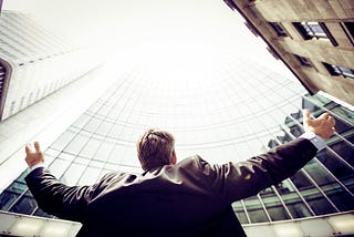 Businessman looking up at tall buildings, arms raised, feeling the joy of acting on his beliefs. | Photo by Razvan Chisu on Unsplash