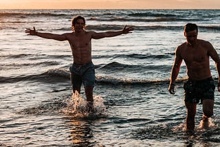 Two men in swim trunks standing in the surf.