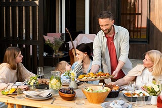 Family eats around a dining table outside, man is passing vegetables