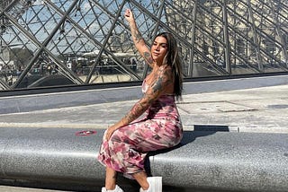 Woman in a pink flowery dress sits on a polished concrete ledge pointing up to the Louvre pyramid.