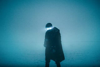 A man in a black coat wearing radiating glasses stands on a snow-covered field at night.