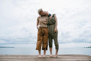 Two women standing on a dock with the water at their back, with one leaning her head on the other’s shoulder.