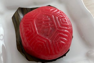 A red and round dumpling sitting on a rectangular piece of banana leaf